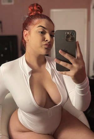 Curvy woman with red hair in white bodysuit taking mirror selfie indoors.