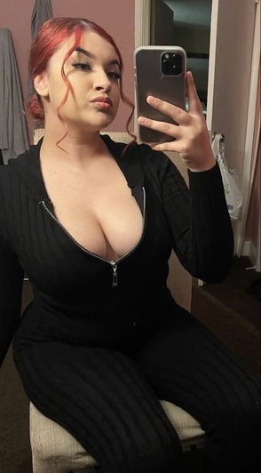 Red-haired woman in black zip-up outfit taking mirror selfie.