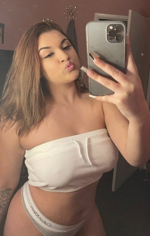 Curvy woman taking mirror selfie in white top and underwear indoors.