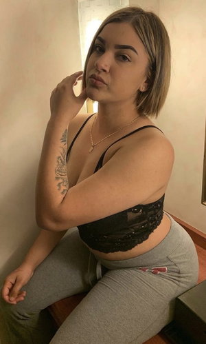 Woman with short hair in black lace top and gray pants, sitting indoors.