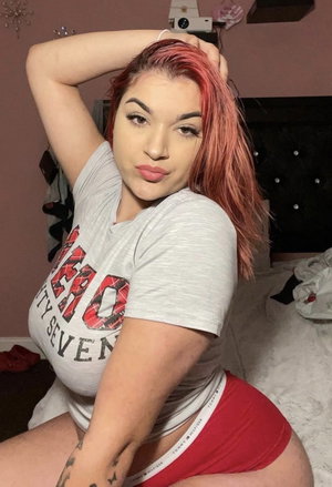 Woman with pink hair in gray t-shirt and red shorts posing on bed indoors.