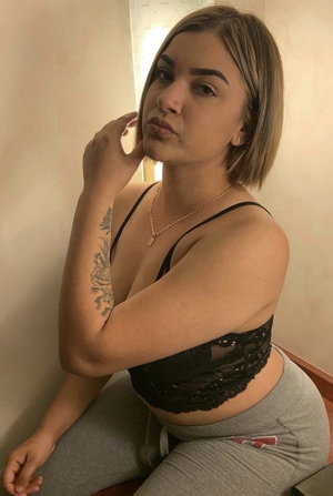 Woman with short hair in black lace top and gray pants, posing indoors near wall.