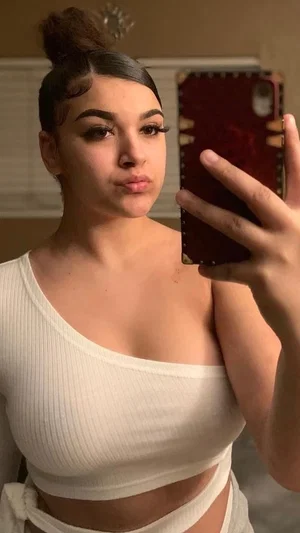 Woman in white ribbed crop top takes mirror selfie, hair in bun, indoor setting.
