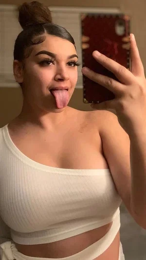 Woman with dark hair in bun, wearing white crop top, sticking tongue out in mirror selfie.