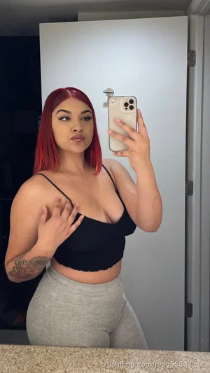 Woman with red hair in black crop top and gray leggings taking mirror selfie indoors.