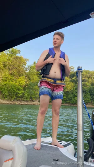 Man in life vest and colorful shorts standing on a boat on a lake.