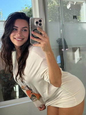 Woman with long dark hair in a white graphic t-shirt, posing in a mirror selfie indoors.