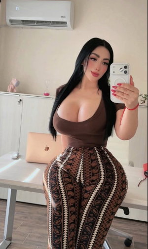 Woman with long black hair in brown top and patterned pants taking mirror selfie in indoor setting.