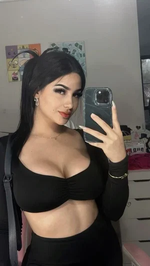Woman with dark hair in black crop top and high-waisted pants, taking mirror selfie in bedroom.