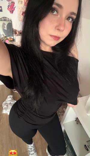 Woman with long black hair in black top and leggings, taking mirror selfie in bedroom.