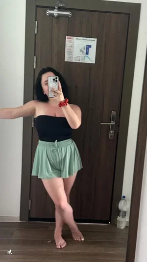 Woman in black strapless top and green shorts poses for mirror selfie in indoor setting.