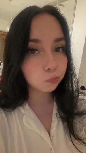 Woman with long dark hair in a white button-up shirt, taking a mirror selfie indoors.