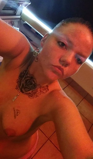 Tattooed woman taking a topless selfie in a kitchen.