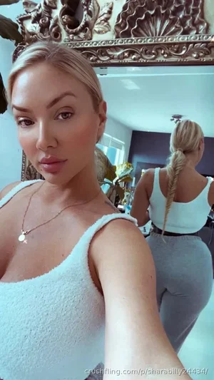 Blonde woman in white top and gray leggings takes mirror selfie in ornate room.