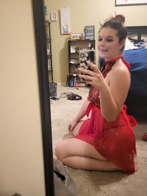 Woman in red lace dress sits on floor, taking mirror selfie in bedroom.