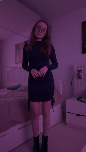 Woman with glasses in black mini dress and boots in bedroom.