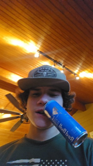 Man with curly hair wearing cap and t-shirt, drinking Red Bull in wooden room.