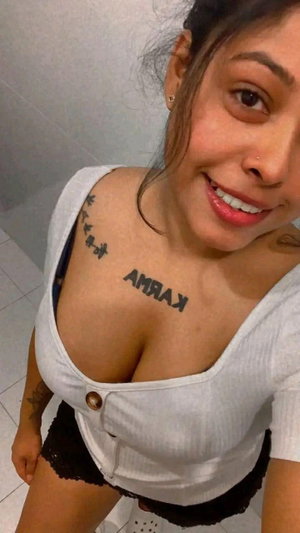 Woman with tattoos in white top taking selfie indoors.