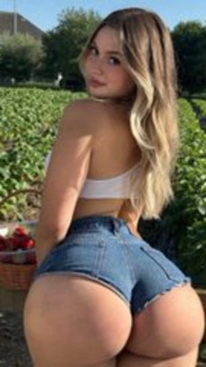 Woman with long blonde hair in white crop top and denim shorts posing outdoors in a strawberry