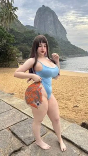Woman in blue swimsuit with large colorful tattoo on hip, standing on stone path near beach with