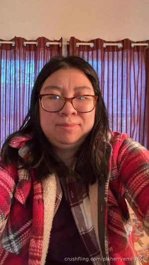 Woman with dark hair and glasses wearing a plaid jacket indoors.