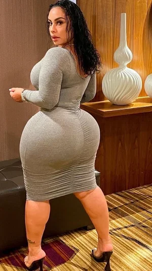 Woman in tight gray dress posing in indoor setting with wooden furniture.