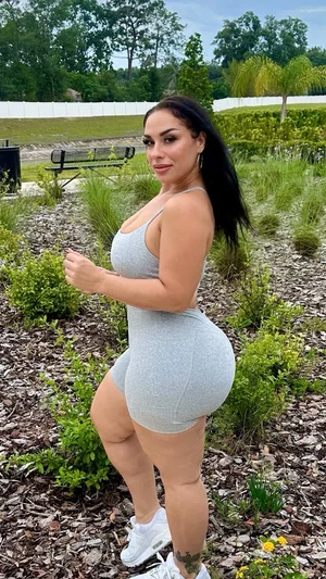 Woman in gray bodysuit posing outdoors in a garden with greenery and mulch.