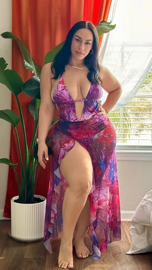 Woman in colorful high-slit dress posing indoors near window and plant.