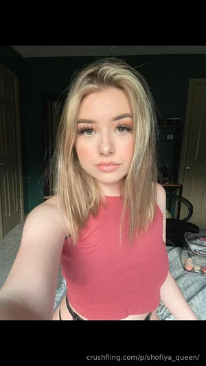 Woman with blonde hair in a red crop top, taking a selfie in a bedroom.