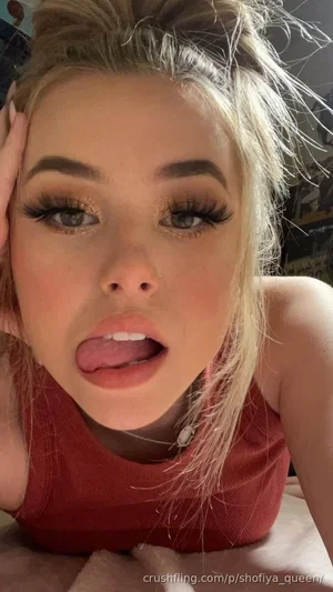 Woman with blonde hair and makeup, wearing a red tank top, lying on a bed.