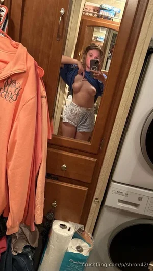 Woman in patterned shorts and open blue shirt takes mirror selfie in laundry room.