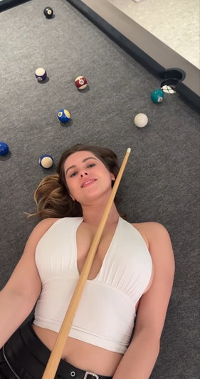 Woman in white halter top lying on pool table holding cue.