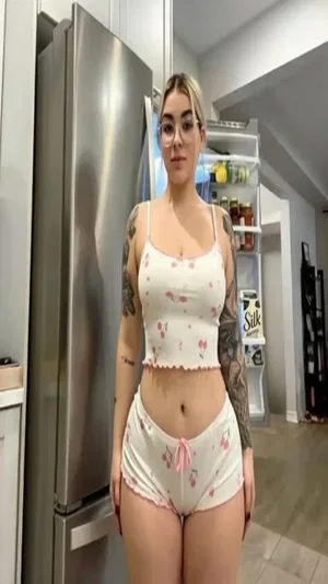 Blonde woman with tattoos and glasses in cherry pajamas in kitchen.