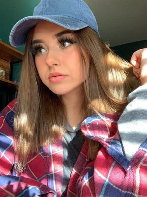 Woman with long brown hair wearing blue cap and plaid shirt indoors.