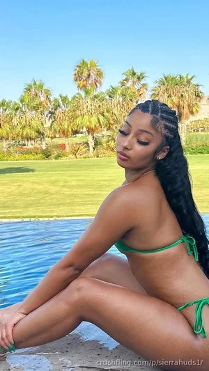 Woman in green bikini sitting by poolside, long braided hair, outdoor setting.