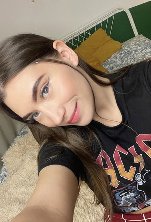 Woman with long brown hair in black graphic tee, smiling in bedroom selfie.
