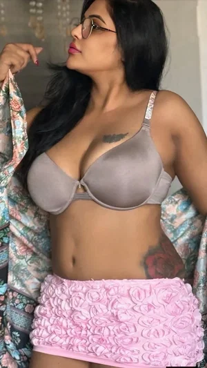 Woman with long black hair in gray bra and pink skirt, holding floral robe, indoors.