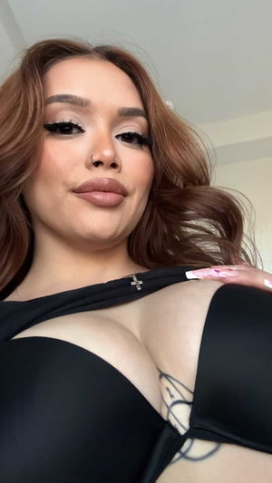 Buxom woman with brown wavy hair in black bra indoors.