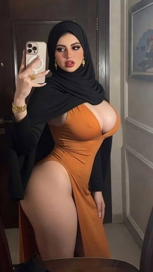 Woman in black hijab and orange dress taking mirror selfie indoors.
