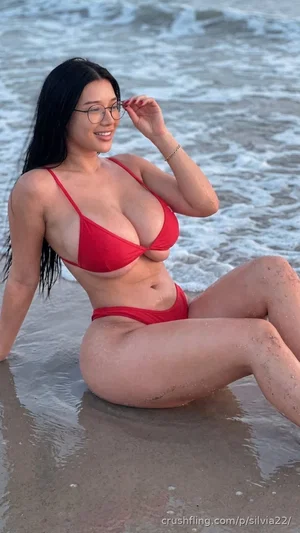 Woman in red bikini sitting in shallow ocean water at beach, smiling.