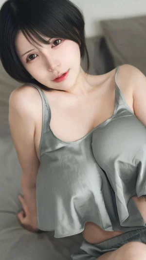 Woman with short black hair in gray satin top, lying on bed indoors.