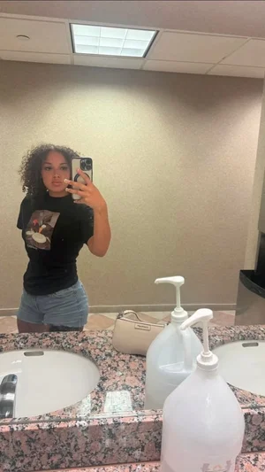 Woman with curly hair in black t-shirt and denim shorts taking mirror selfie in bathroom.