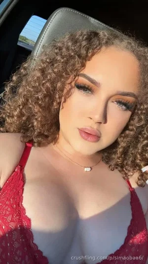 Woman with curly hair in red lace top sitting in car, wearing necklace.