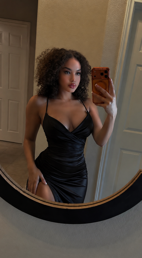 Woman with curly hair in black dress taking mirror selfie indoors.