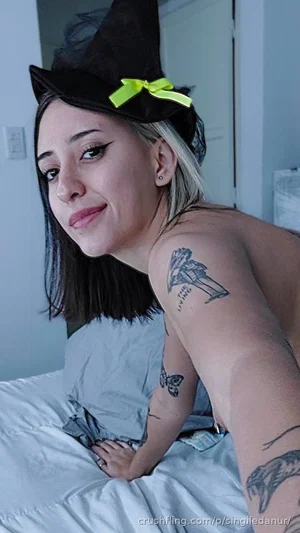 Woman with tattoos and witch hat, topless, sitting on bed indoors.