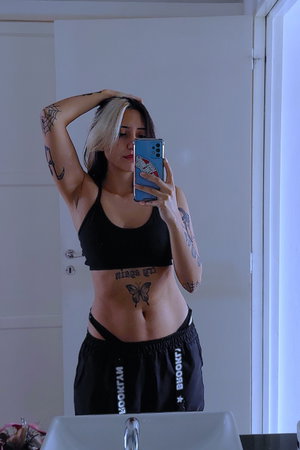Woman with tattoos in black crop top and sweatpants takes mirror selfie in bathroom.