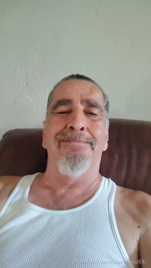 Man with gray beard and mustache wearing white tank top, lying on brown couch indoors.