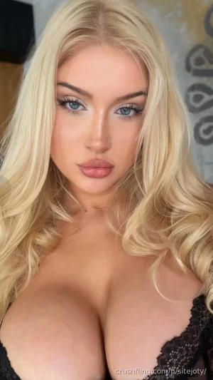 Blonde woman in black lace top, close-up selfie, indoor setting.