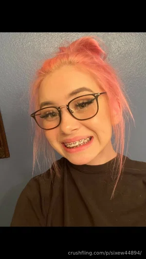 Woman with pink hair and glasses, wearing a black turtleneck, smiling indoors.