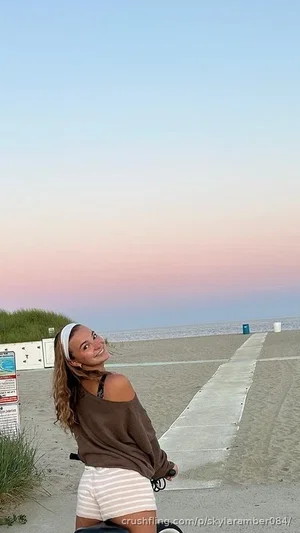 Woman in brown off-shoulder top and striped shorts on beach, smiling at camera.